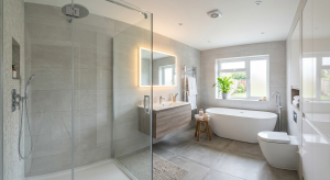 A modern redesigned bathroom in a UK home featuring large-format grey wall tiles, a glass walk-in shower, a floating wooden vanity with an LED mirror, and a freestanding bathtub by the window.